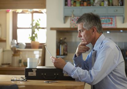 A man in his 50s sits at the kitchen table, looking thoughtfully at his laptop, which rests on his briefcase. 