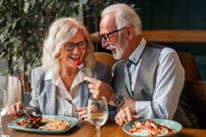 Elegant elderly man and woman in formal attire smiling and talking warmly while sharing a meal and wine together at a cozy restaurant. Senior man in a suit feeding his wife in a restaurant.