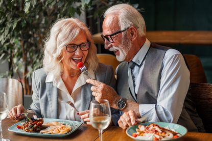 Elegant elderly man and woman in formal attire smiling and talking warmly while sharing a meal and wine together at a cozy restaurant. Senior man in a suit feeding his wife in a restaurant.