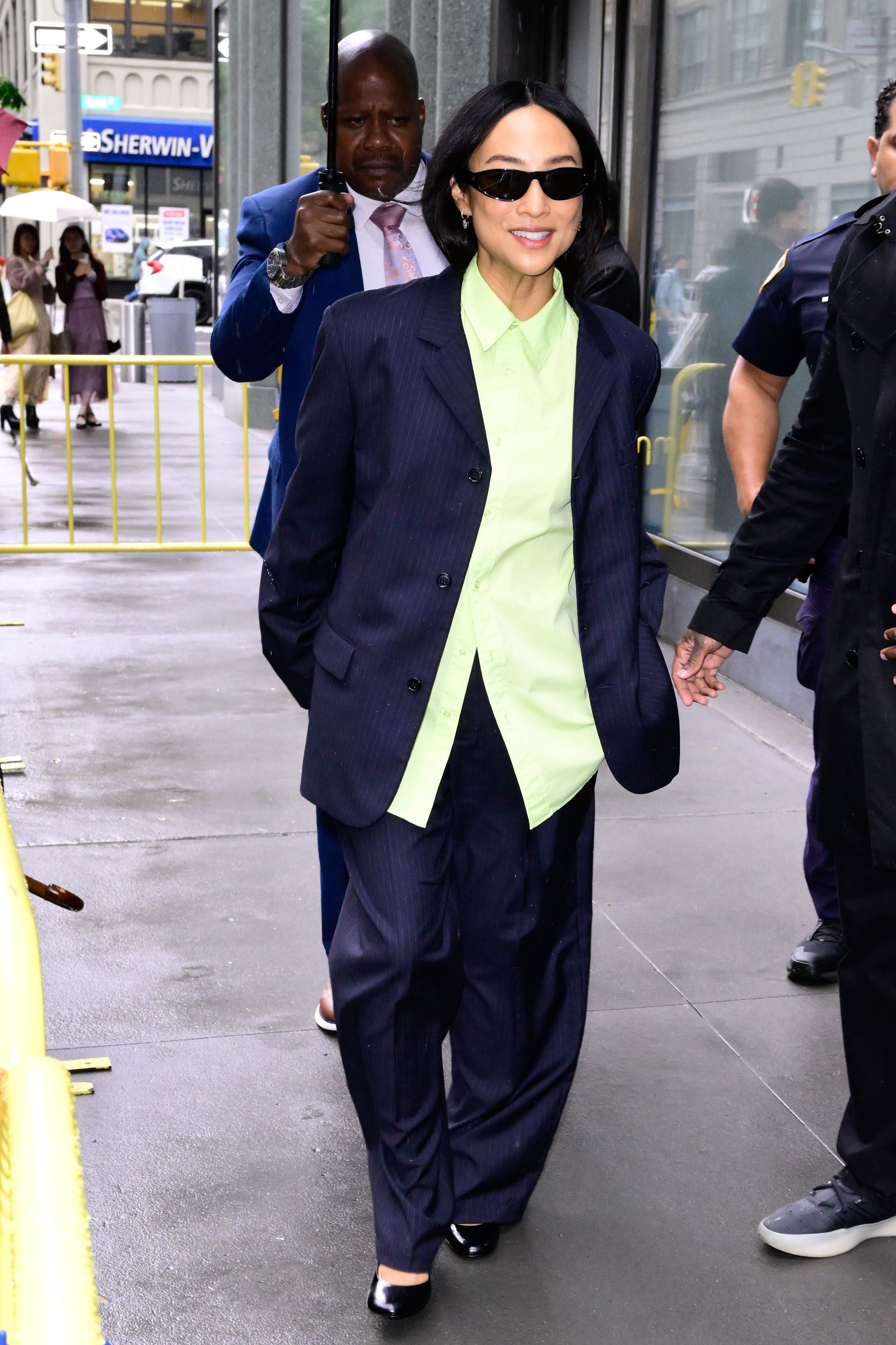 Greta Lee wearing a navy blue pinstripe blazer and pants, lime green button-down, and black flats