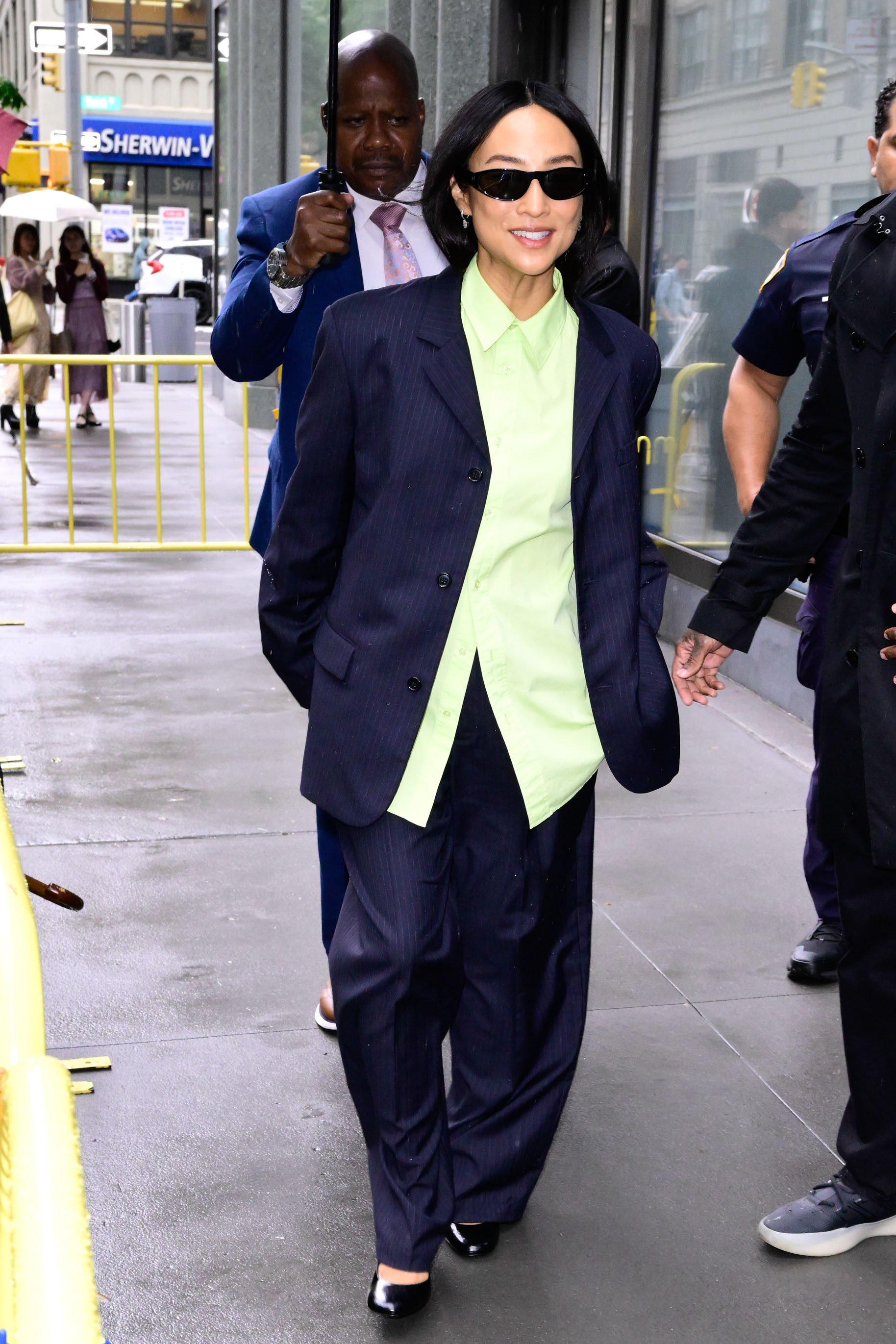Greta Lee wearing a navy blue pinstripe blazer and pants, lime green button-down, and black flats