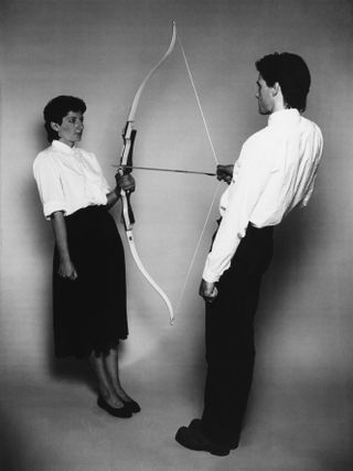 Marina Abramovic stands leaning on the wall holding the raiser of a bow, while her former partner holds a strung arrow towards her. The photo is black & white.