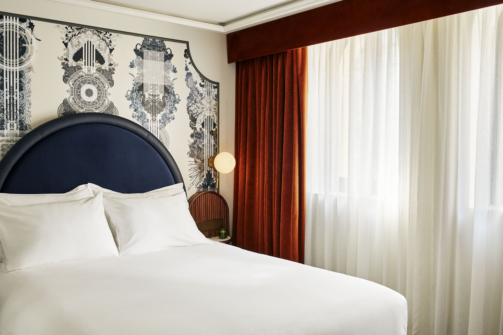 This is an image of a hotel bedroom with an all-white bed and a curved navy blue headboard. Behind the bed, there is navy-printed wallpaper. The wall to the right has white and burnt orange curtains.