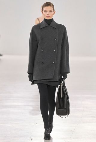 A model from Max Mara's fall runway wearing a gray pea coat over a black turtleneck and gray miniskirt styled with black tights, black shoes, and a black bag.