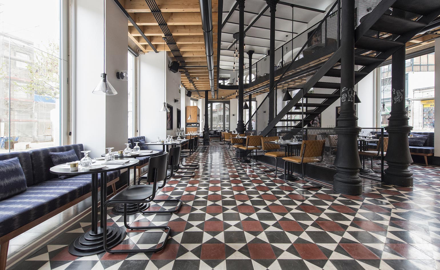 Interior view of the 1908 bar area. To the left, in front of large floor-to-ceiling windows is a wide sitting area in deep blue, with metal black tables and chairs across. Geometric tiles in red, black, and white cover the floor, while black metal stairs lead to the second floor.