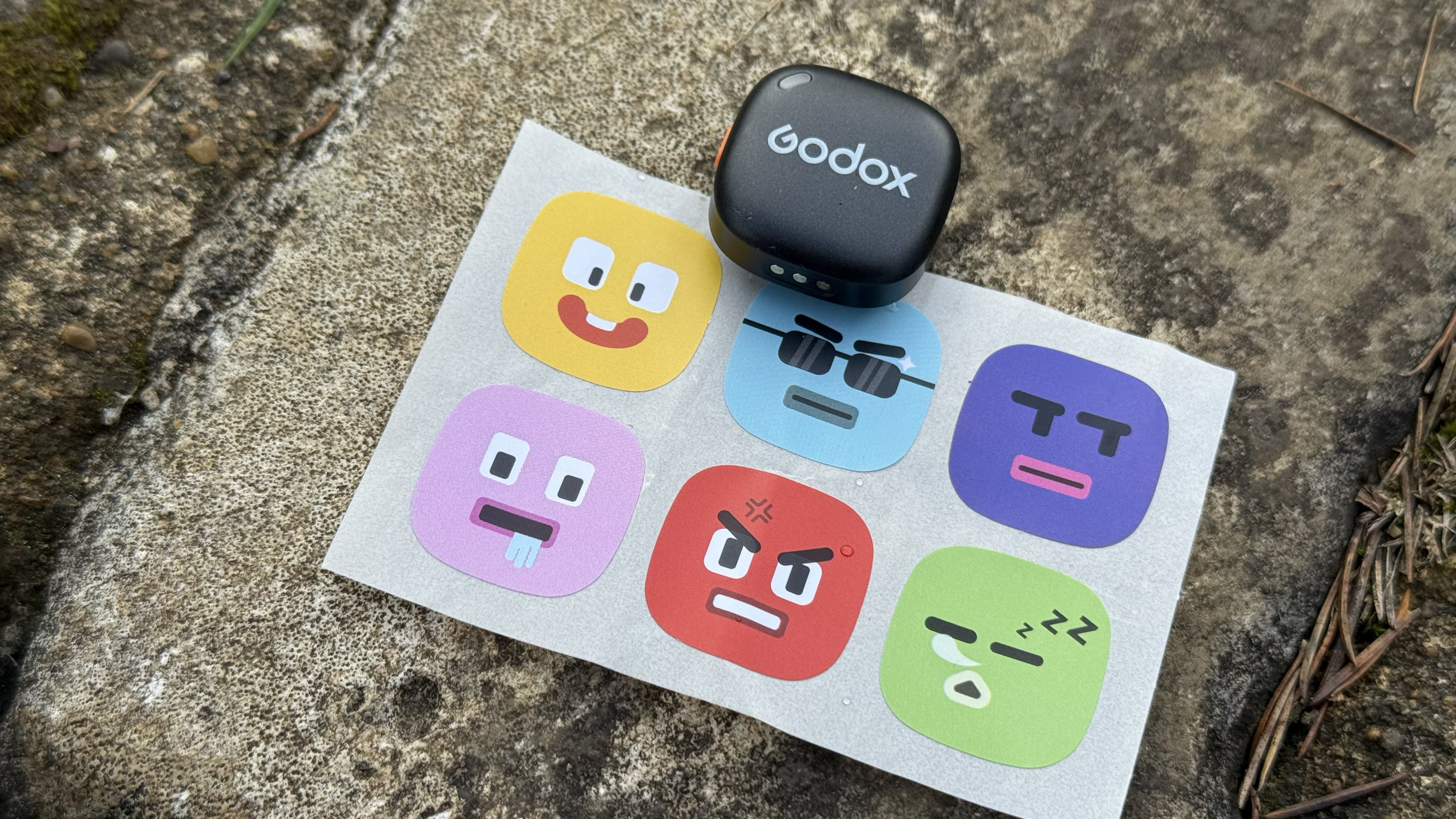 Shot of a wireless transmitter branded with the Godox logo. It sits next to a sheet of &amp;lsquo;cute&amp;rsquo; smiley style stickers that can be used to personalise the transmitter.