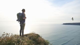 A male hiker stands carrying his backpack looking out to sea from an Atlantic clifftop as a seagull flies past