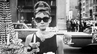 Audrey Hepburn as Holly Golightly, holds a cup and a paper bag while looking into one of the window displays at Tiffany's in a still from the film, 'Breakfast at Tiffany's,' directed by Blake Edwards. She wears sunglasses, a little black dress, long gloves and a tiara in her chignon. (Photo by Paramount Pictures/Getty Images