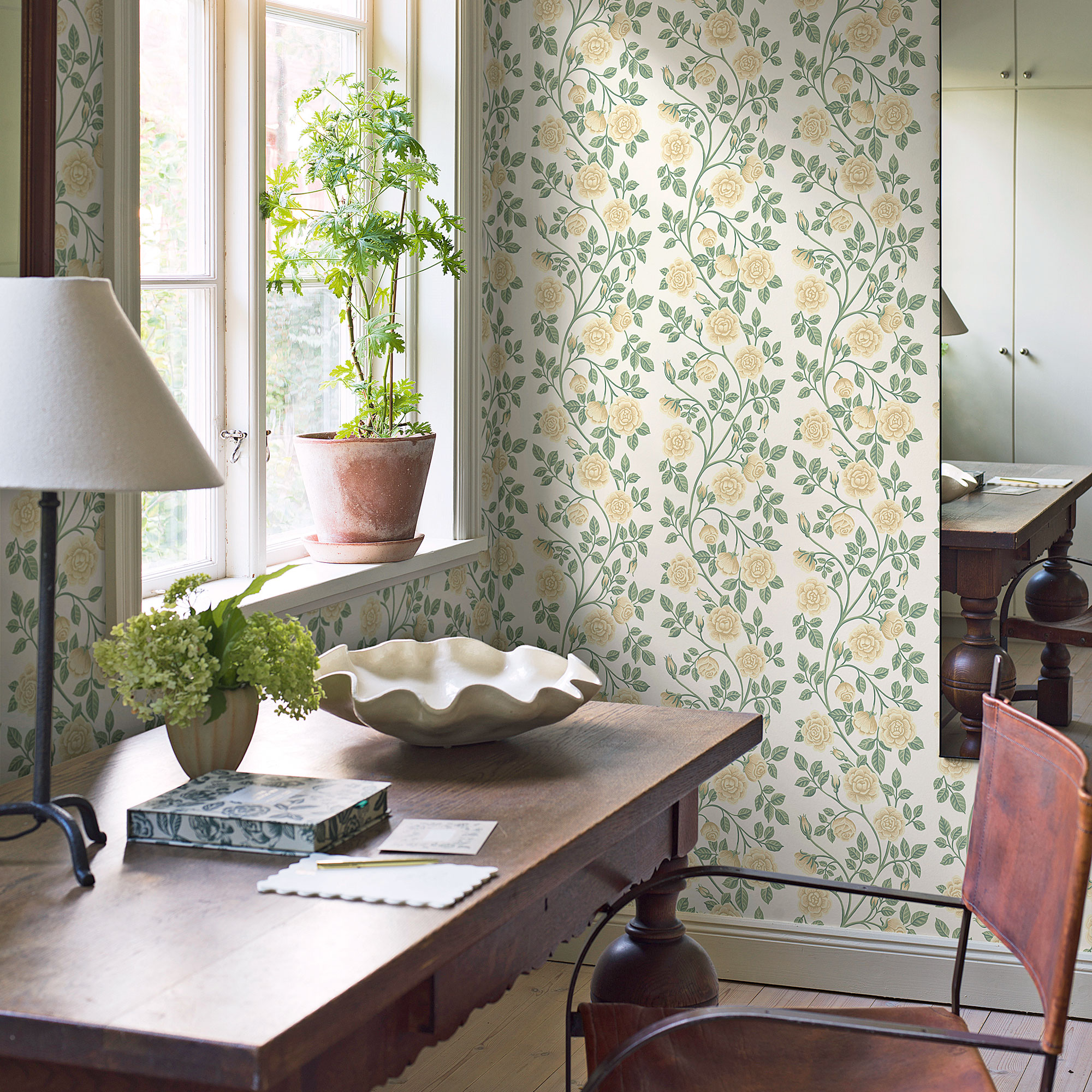 Green floral wallpaper in a study with wooden desk and a plant in terracotta plant pot
