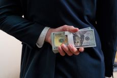 A man wearing a suit holds a stack of cash behind his back.