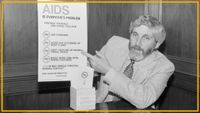 A black and white photo shows a man with long hair and a beard wearing a suit and patterned tie pointing at a poster labeled "AIDS is everyone's problem"
