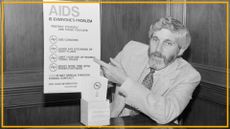 A black and white photo shows a man with long hair and a beard wearing a suit and patterned tie pointing at a poster labeled "AIDS is everyone's problem"
