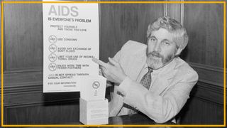 A black and white photo shows a man with long hair and a beard wearing a suit and patterned tie pointing at a poster labeled "AIDS is everyone's problem"