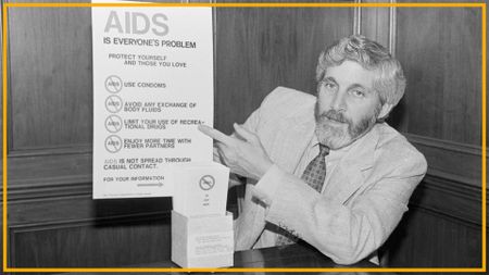 A black and white photo shows a man with long hair and a beard wearing a suit and patterned tie pointing at a poster labeled "AIDS is everyone's problem"