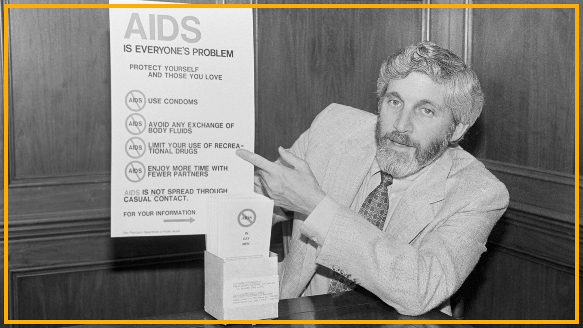 A black and white photo shows a man with long hair and a beard wearing a suit and patterned tie pointing at a poster labeled "AIDS is everyone's problem"