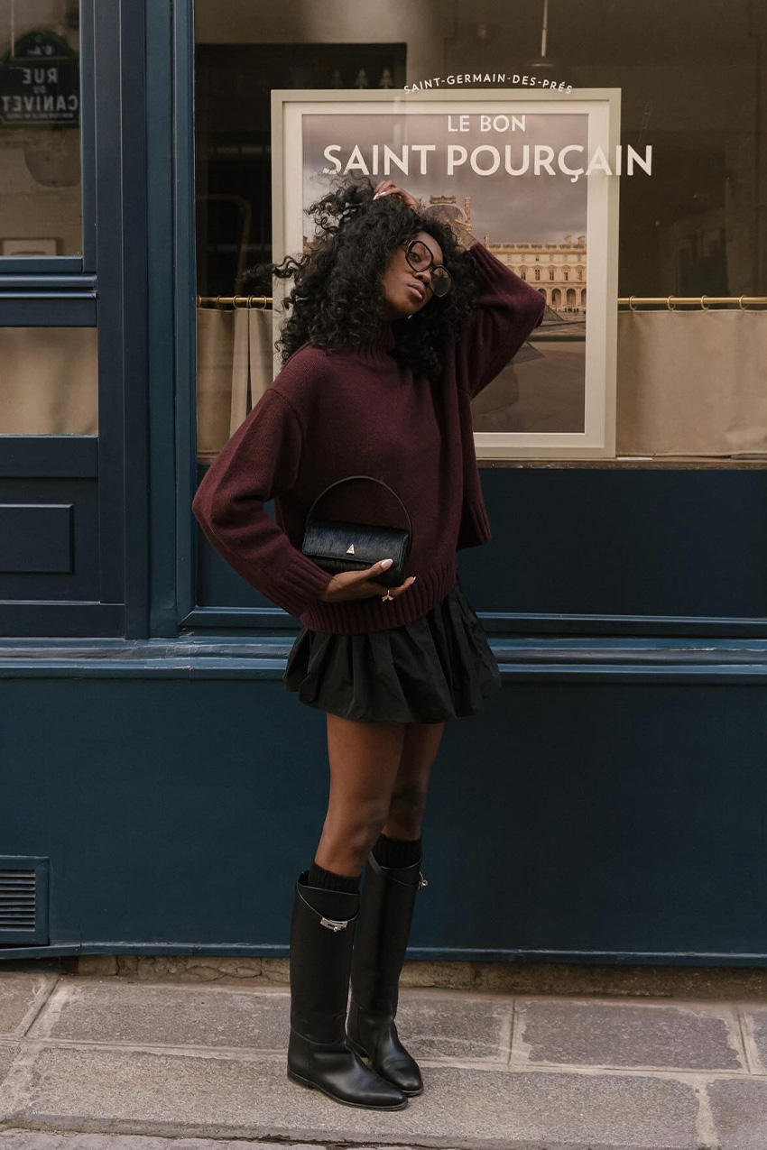 Fall shoe trends in Paris are shown in a photo of a woman standing on the sidewalk wearing the utilitarian riding boots trend with a red turtleneck sweater, black ballon mini skirt, and black handbag