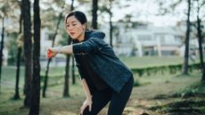 Asian woman wearing a grey hoodie and black leggings resting after working out outdoors. Checking pulse and training performance on fitness app on her smartwatch.