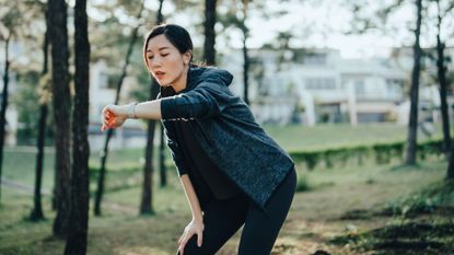 Asian woman wearing a grey hoodie and black leggings resting after working out outdoors. Checking pulse and training performance on fitness app on her smartwatch.