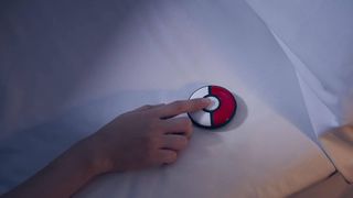 Promo image of the Pokémon Go Plus accessory on a pillow, with someone touching its central button.