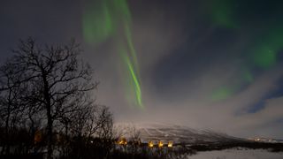 northern lights appear as thin ribbons of green light in a partly cloudy sky. Below are the lights outside cabins at Abisko Turiststation.