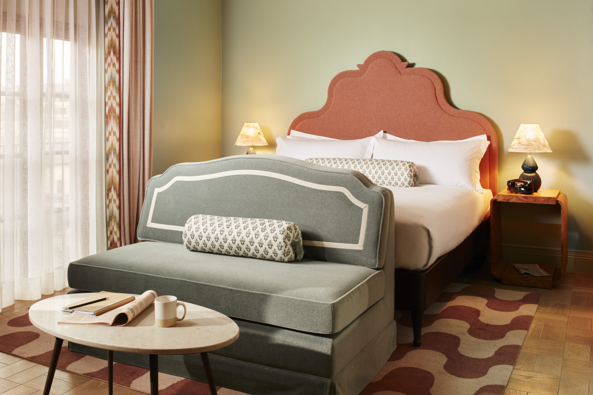 A quirkily decorated, retro-inspired hotel room with a patterned bordeaux and cream rug, tiled floors in light brown, a terracotta-hued headboard in a sculptural decor, and a teal blue bench at the feet of the bed.