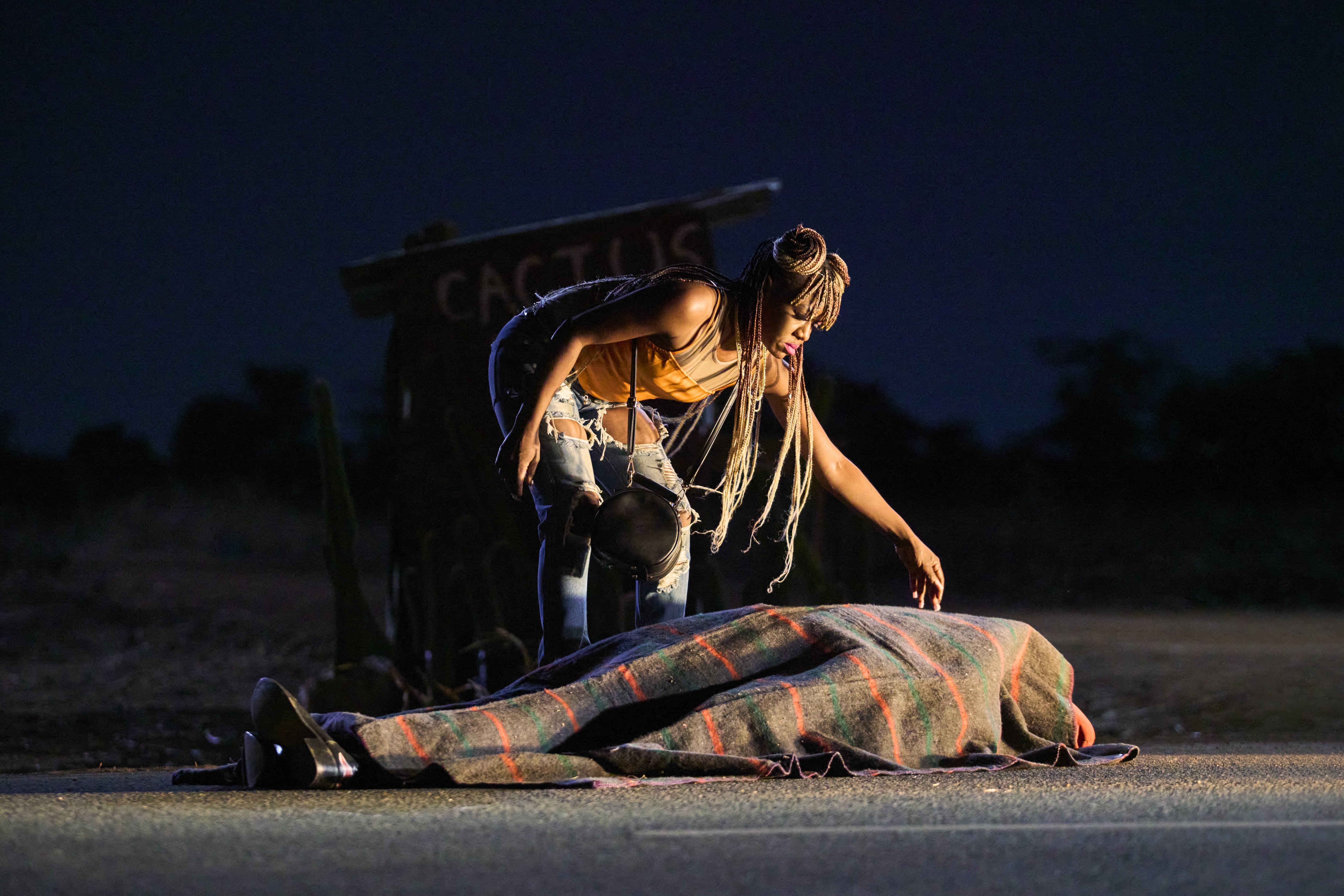 A woman (Elizabeth Chisela as Nsansa) touches a dead body laying in the road, covered with a blanket, in 'On Becoming a Guinea Fowl.'