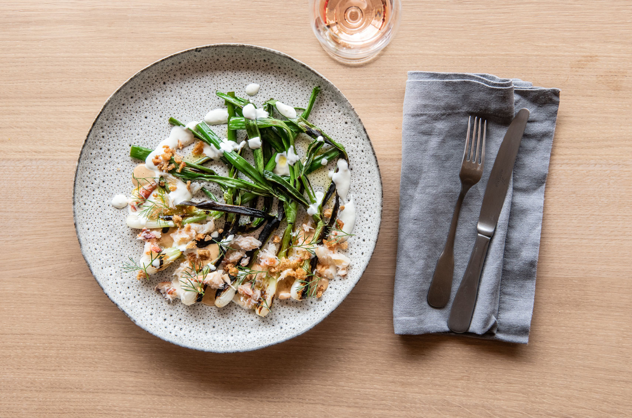 A plate of charred spring onion and crab on a table with a glass of rose and cutlery
