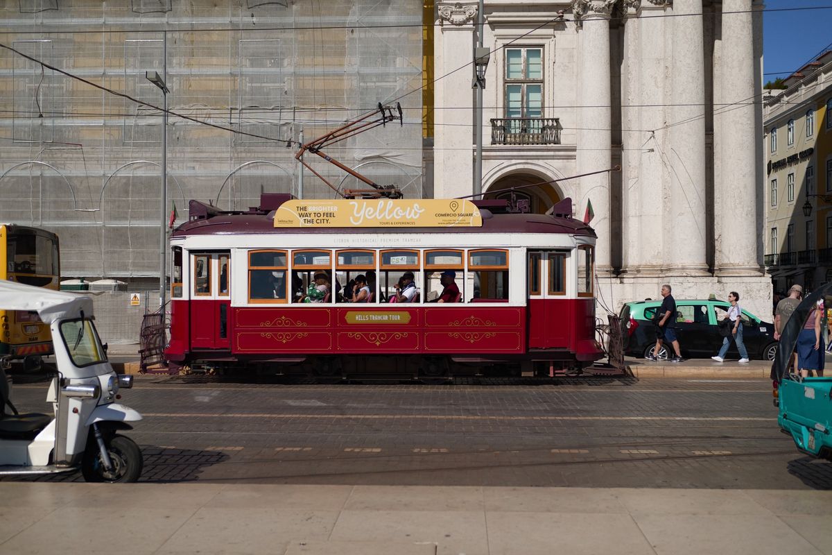 Red vintage sightseeing tram carrying passengers through Pra&ccedil;a do Com&eacute;rcio, with tuk-tuks and pedestrians nearby.
