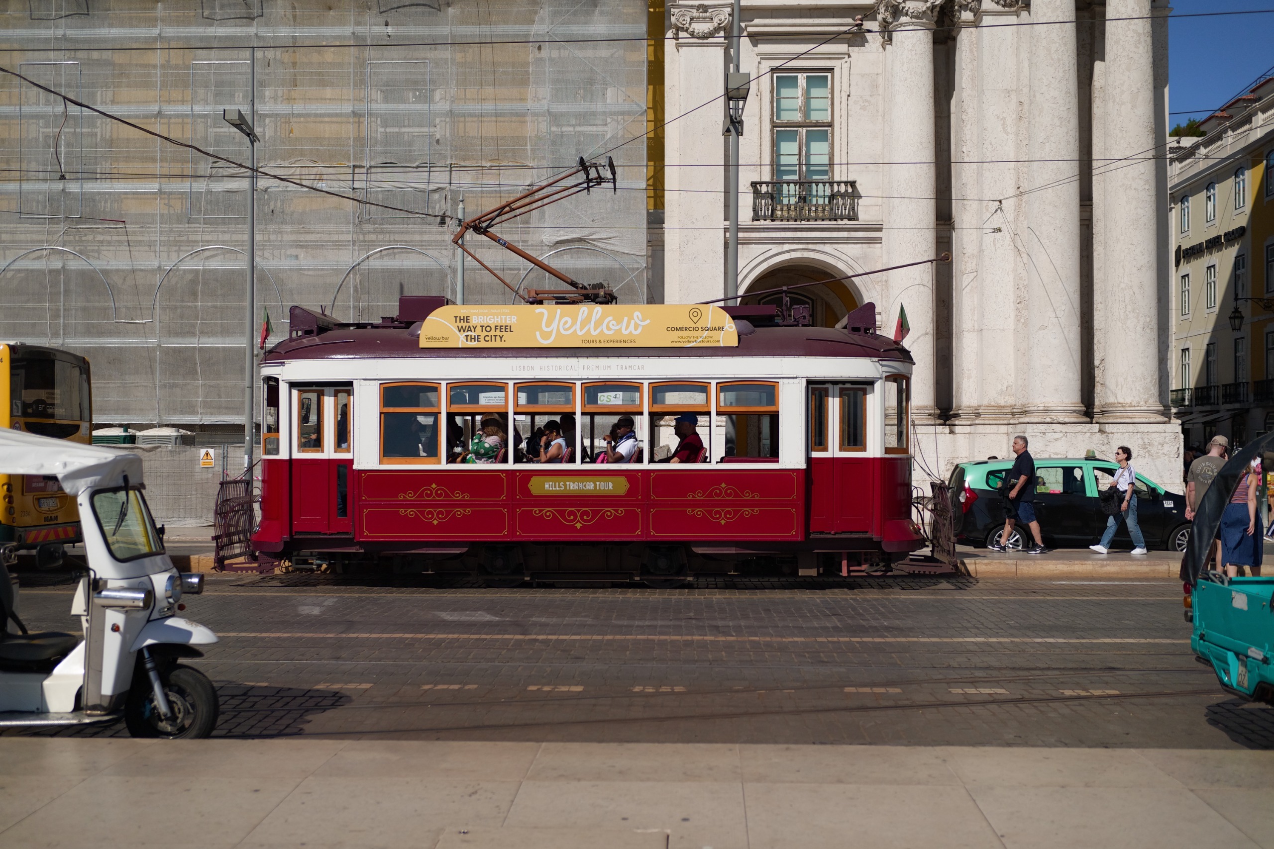 Red vintage sightseeing tram carrying passengers through Pra&amp;ccedil;a do Com&amp;eacute;rcio, with tuk-tuks and pedestrians nearby.