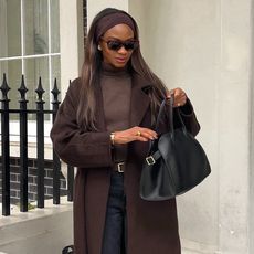 British style influencer Lorna Symphony poses in black sunglasses, a brown headband, brown turtleneck sweater, brown coat, black The Row Margaux bag, and black jeans