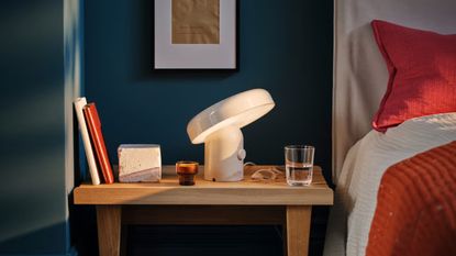 Image of a dark blue bedroom with a wite bed with red and white bedding. There is a wooden side table that is in the center of the frame that has a white Bluetooth speaker lamp on it, as well as a water glass and some books.