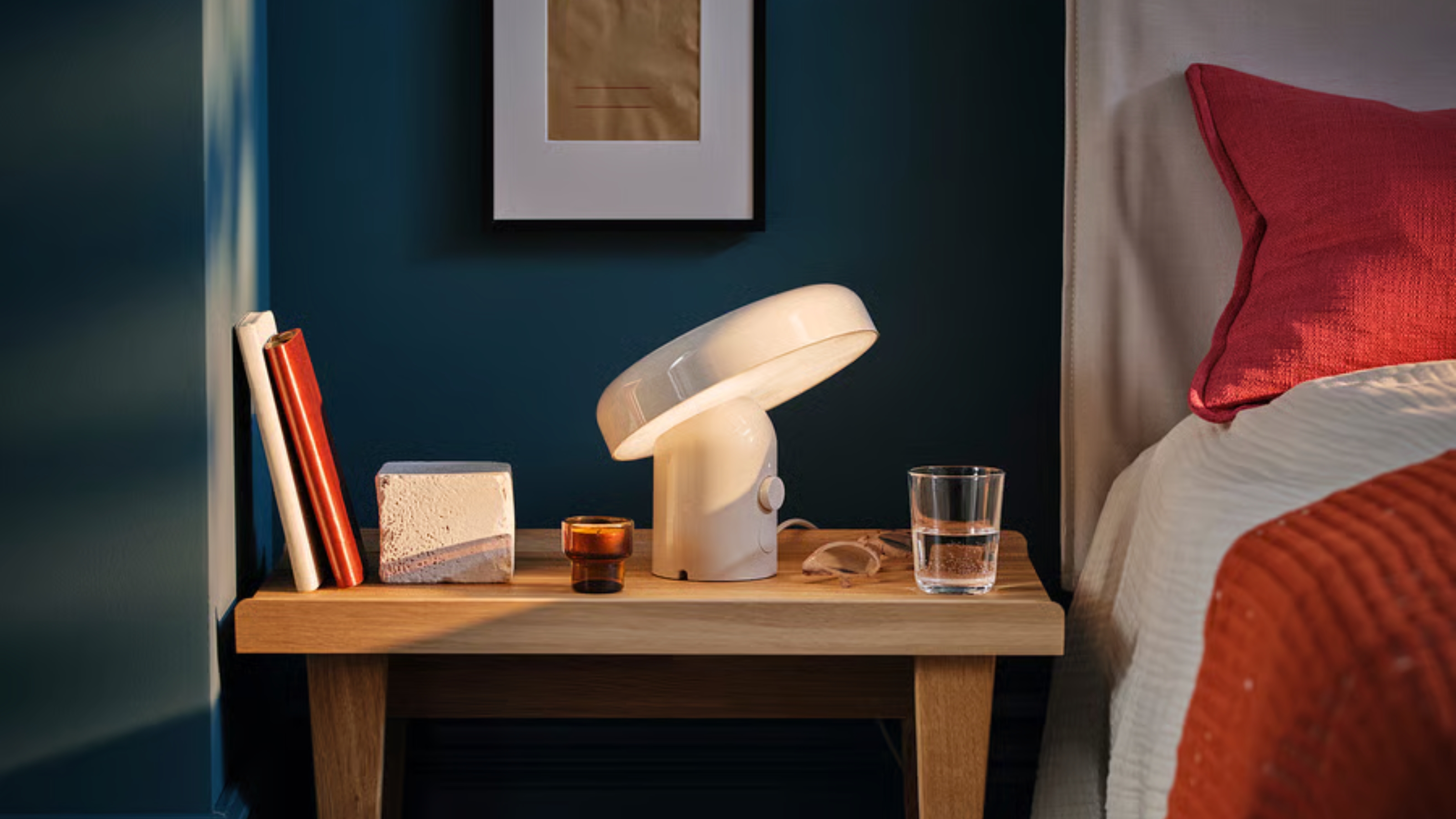 Image of a dark blue bedroom with a wite bed with red and white bedding. There is a wooden side table that is in the center of the frame that has a white Bluetooth speaker lamp on it, as well as a water glass and some books. 