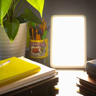 Rectangular sad lamp on a desk next to a pile of notebooks and pot of pens