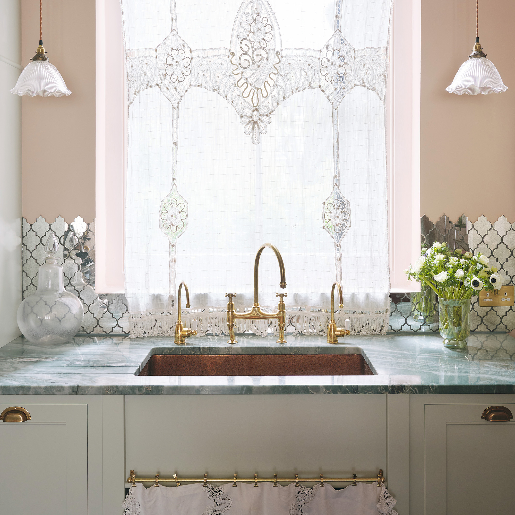 lace curtains on kitchen window in front of brass taps and brass inset sink with cream cabinets and grey marble worktops devol