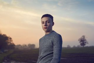 Sam Hall as Emmerdale character Samson Dingle, standing in a field with a sunset behind him and wearing a grey jumper. 