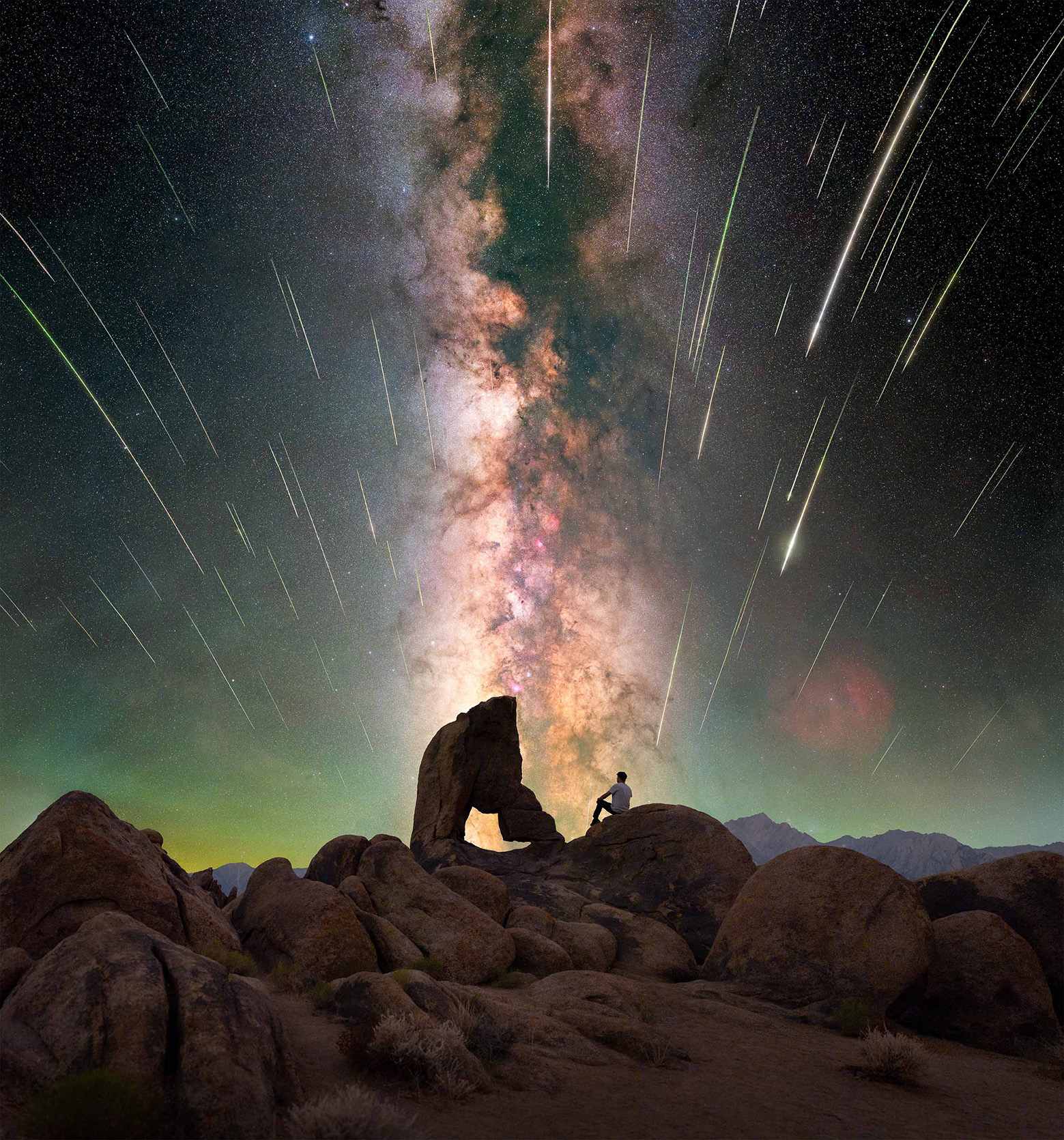 A person sits on a rocky outcrop under a vibrant Milky Way, with swirling stars and meteor trails illuminating the night sky