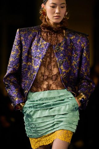 A model in Saint Laurent's Spring 2025 show wearing a brown lace top, purple jacket, and blue ruffle skirt with yellow lace trim.