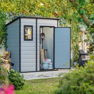 Compact grey garden shed with a pitched roof on a patio against a wall covered in leaves