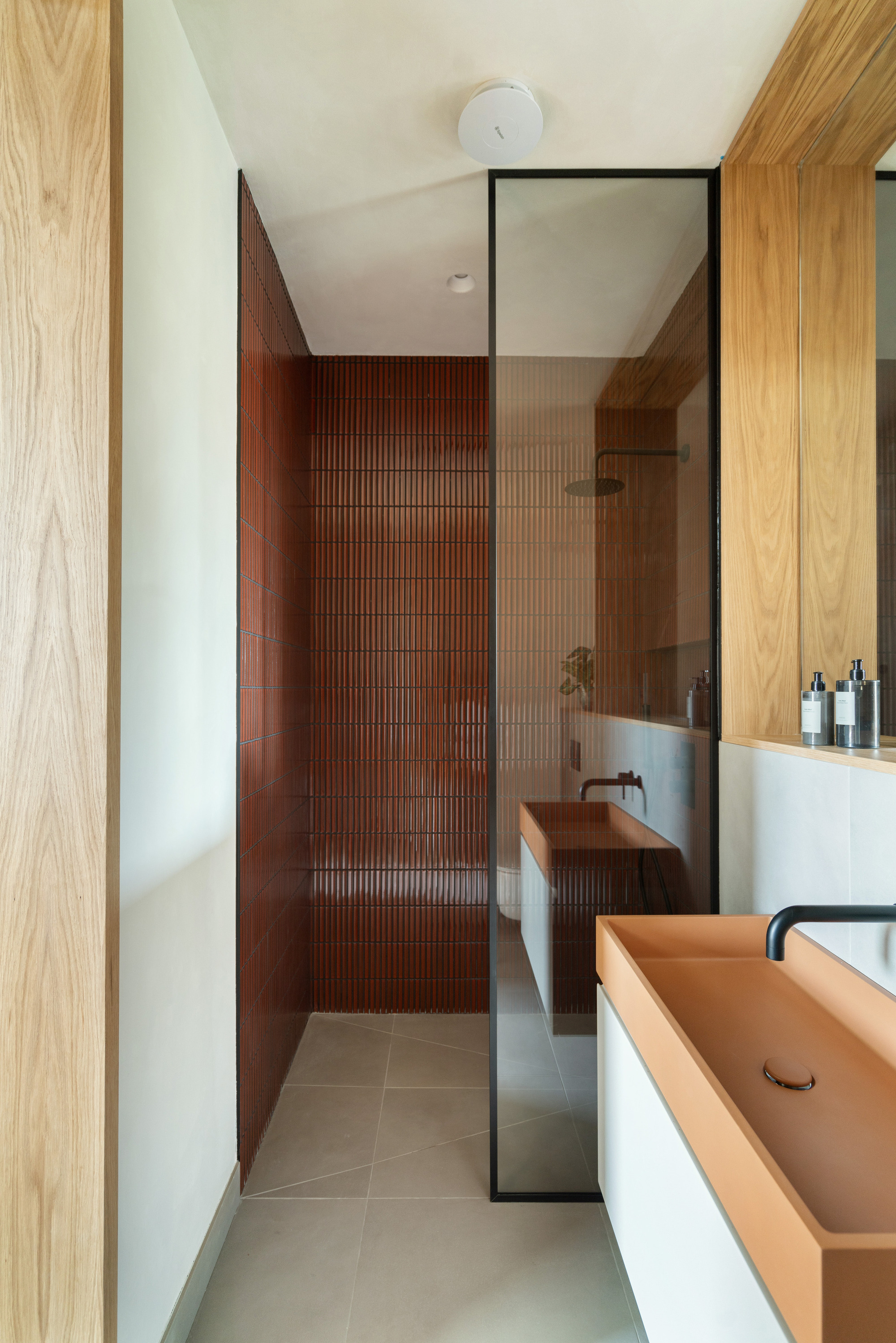 modern bathroom with red kit kat tiles, timber deatures, white walls, gray large format floor tiles, and terracotta-colored sink with black tapware