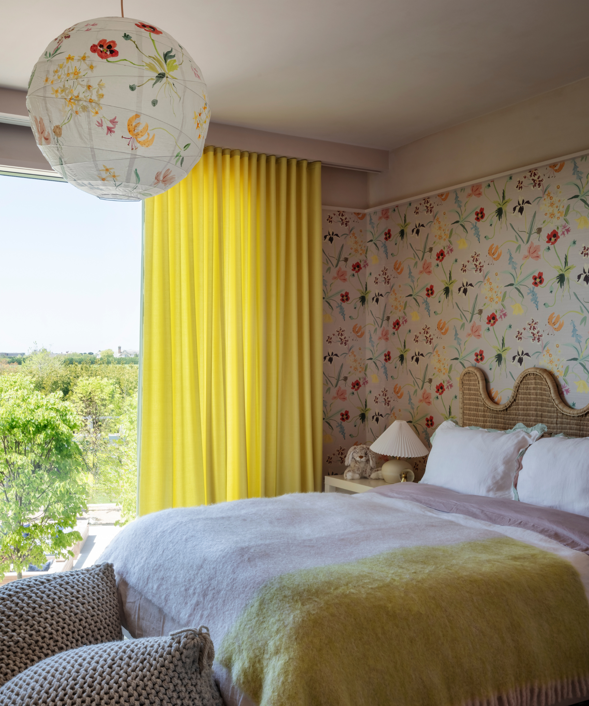 a child&#039;s bedroom with yellow curtains, floral wallpaper, a floral paper ceiling lamp, and a pink and yellow blanket on the bed