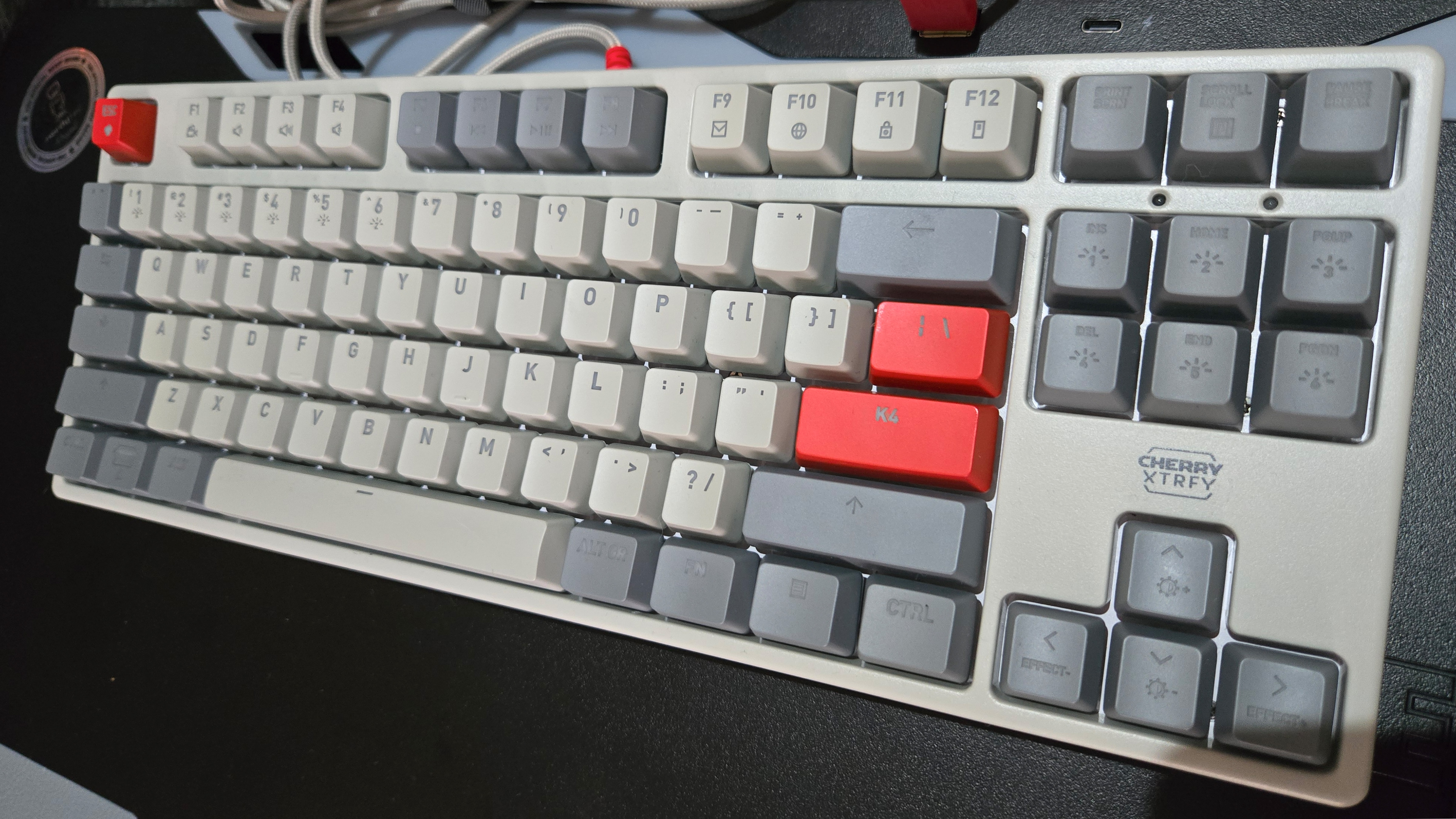 The Cherry Xtrfy K4V2 TKL gaming keyboard in grey with red accent keys. It sits on a black desk surface and is seen at a slight angle. The RGB lights are off.