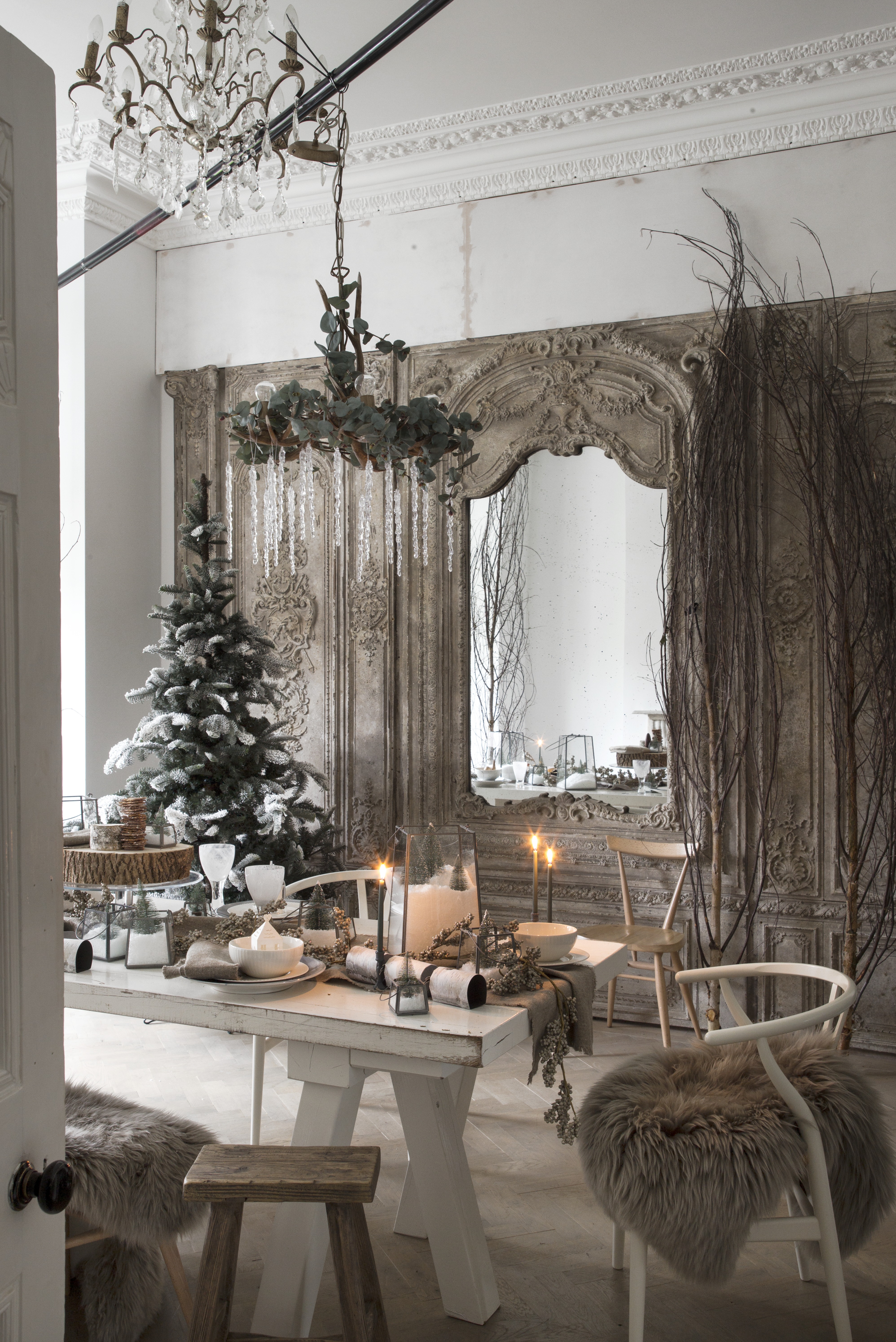 A Christmas dining room with a Christmas table decorated in-keeping with the theme of the room: white, natural, and cozy with foliage, lit candles, throw blankets, with the Christmas tree in the corner of the room and a hanging wreath above the table