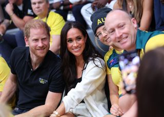 Prince Harry wears a black polo shirt featuring a small Invictus Games logo while he poses for a photo with his wife, Meghan Markle, who wears a white shirt