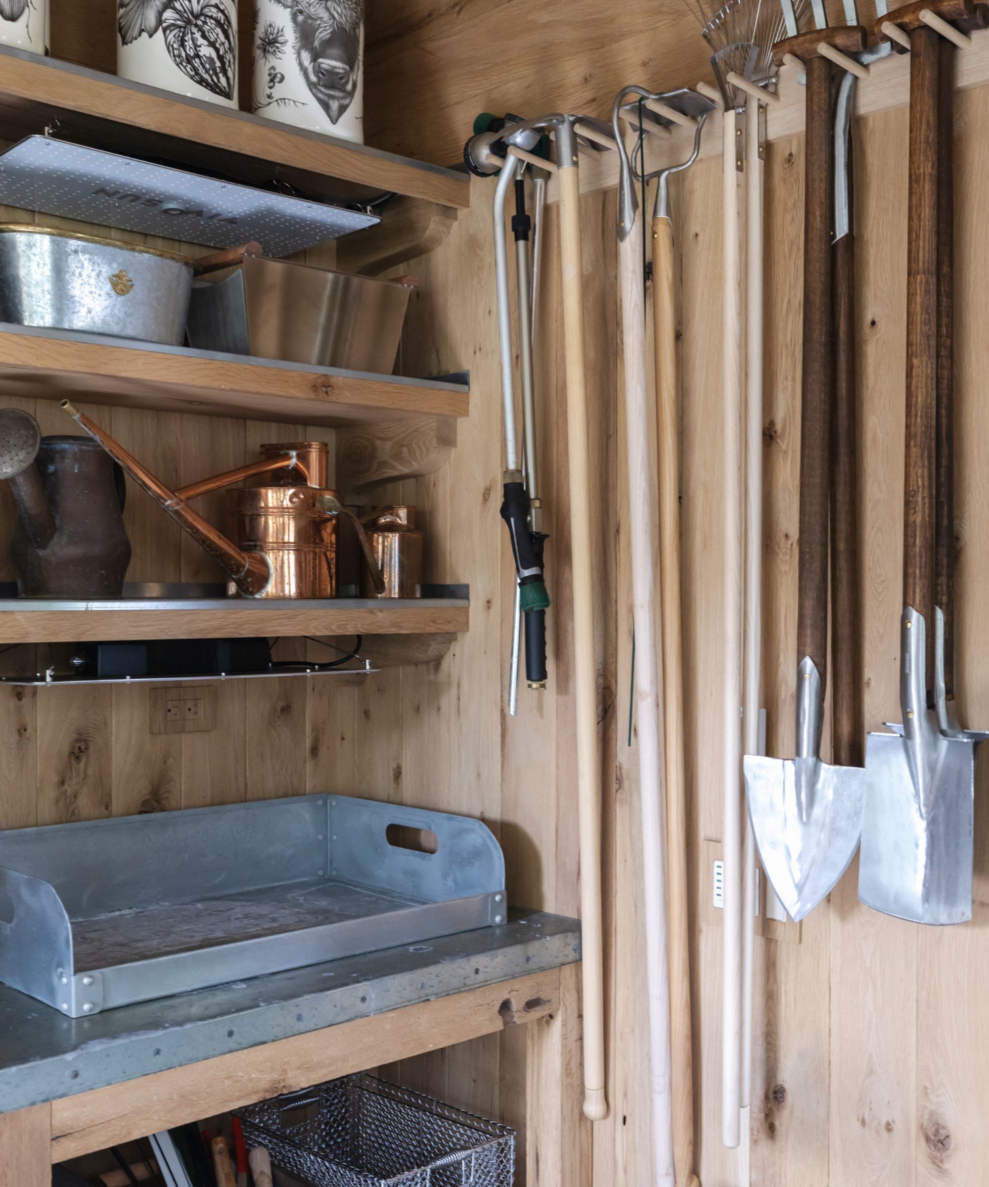 A wall mounted hanging rack with shovels and trowels hanging from it, in a wooden shed. Next to wooden shelving.