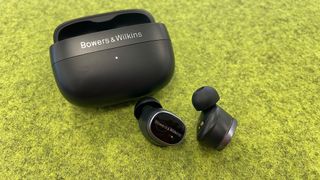 A black pair of Bowers & Wilkins Pi8 wireless earbuds with their charging case on a green surface.