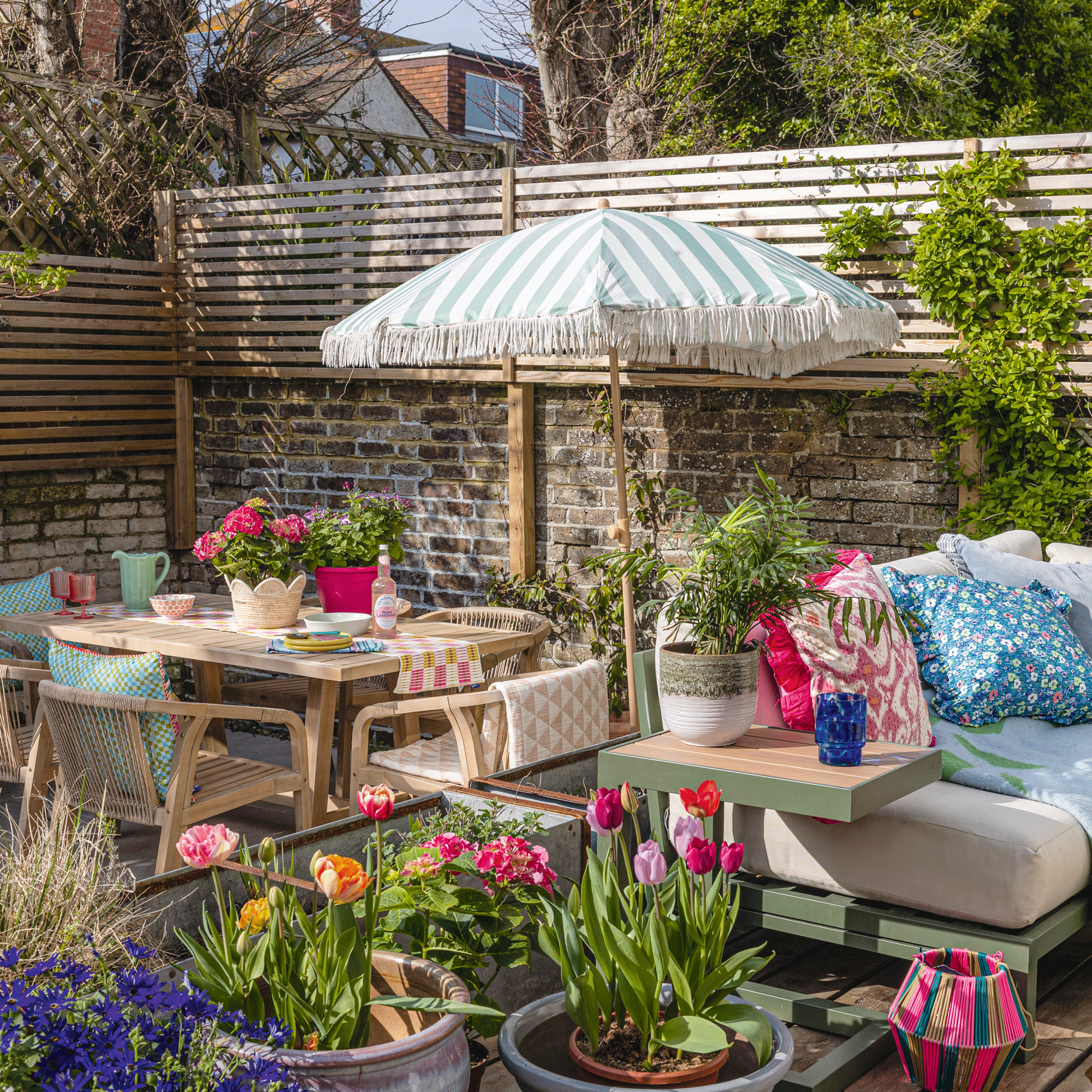 a colourful garden with tulips and two separate decked areas with a dining table and chairs in one and a sofa in the other with a parasol between them