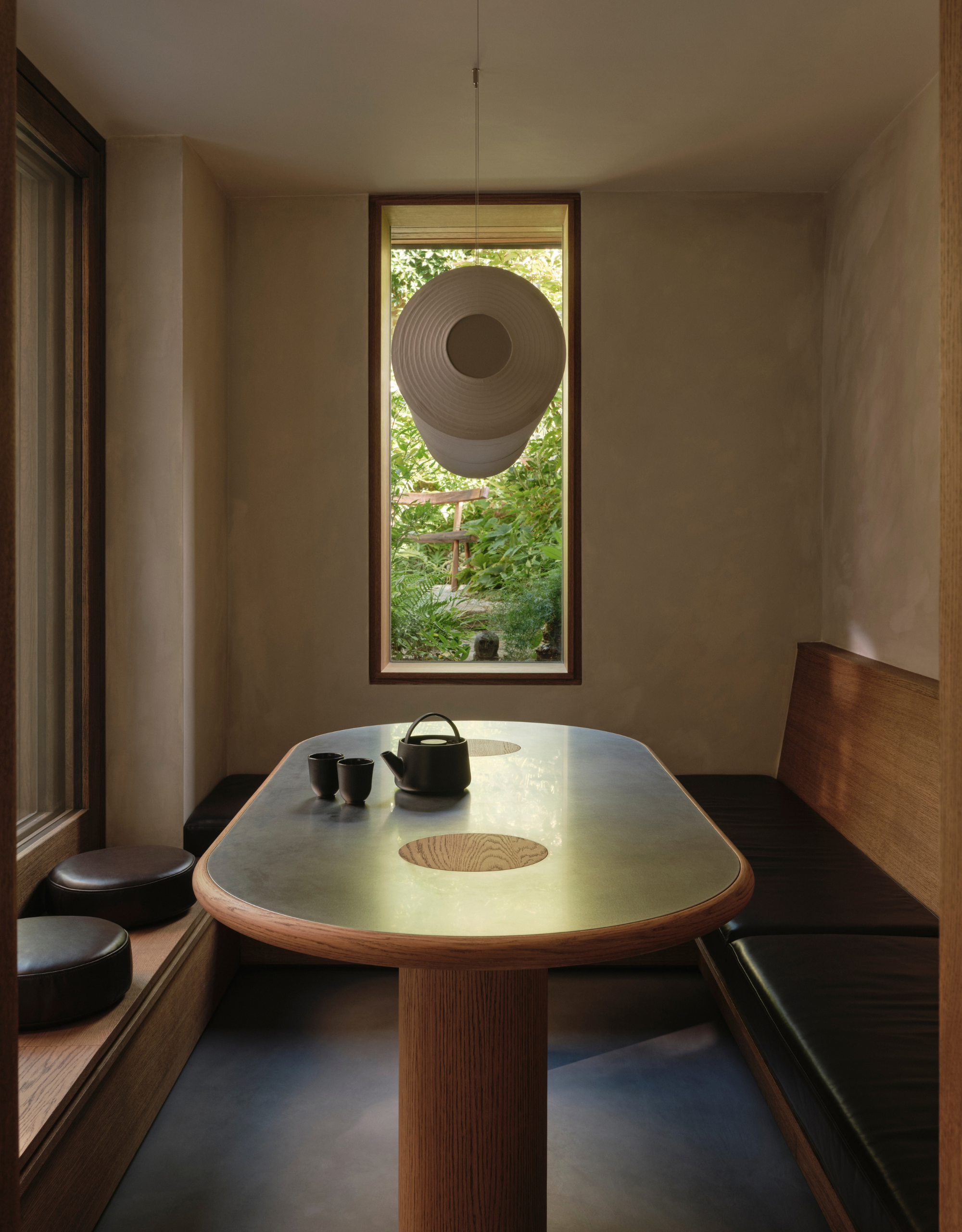 A Japanese dining room with banquette seating, a wood and steel table, a tea set, a window, and a large hanging pendant