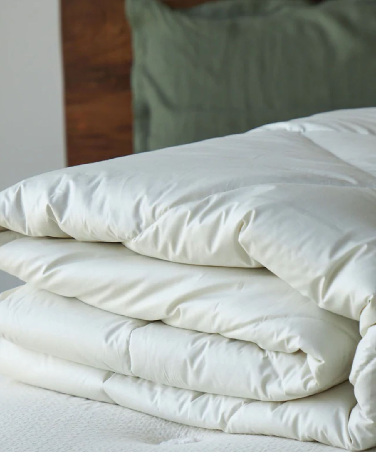 A white folded Avocado Alpaca duvet insert pictured on a white mattress, with a pillow in a green pillowcase against a wooden headboard, beside a white wall, behind.