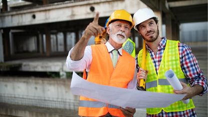 An older construction worker mentors a younger worker on a construction site.