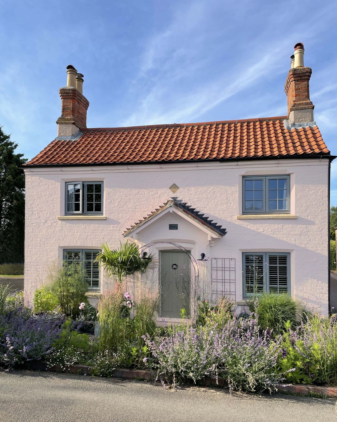 Image of the exterior of a heritage home that is painted a soft pink with soft sage trim and doors. There is a wild garden in the front of the house.
